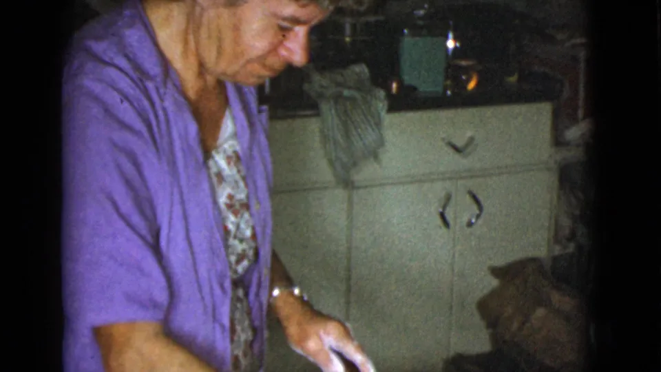 A woman in a purple cardigan and floral apron looks down while working at a kitchen counter in the 1968 home movie.