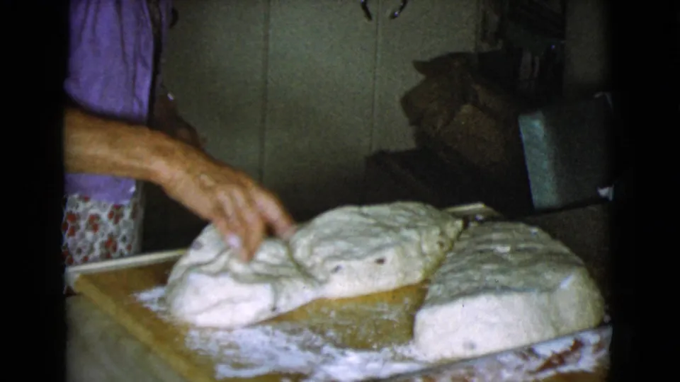 A woman in a purple top works a dough section on a floured wooden board, with cut dough portions visible in the kitchen.