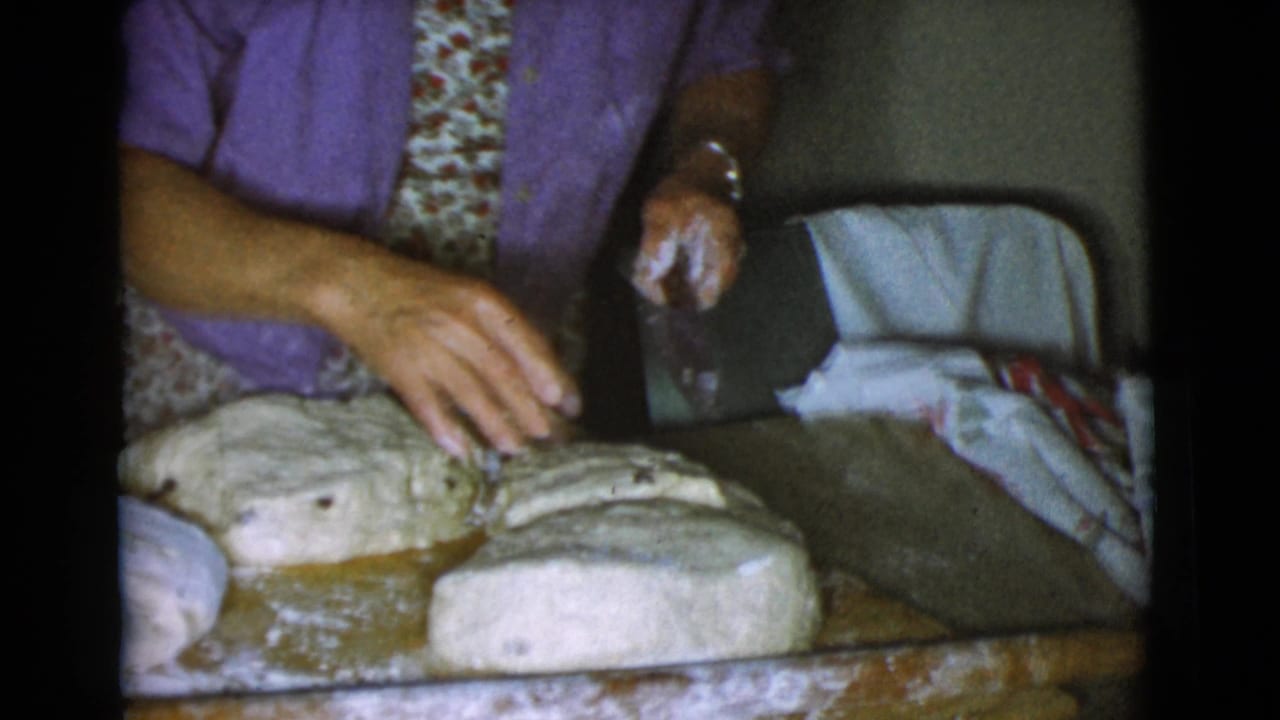 Elder woman reaches over several floured dough portions on a wooden kitchen surface.