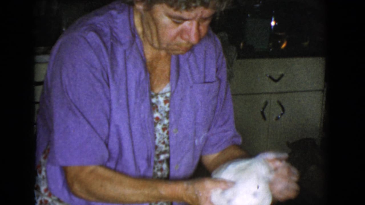 Woman in a purple shirt kneading a mound of dough in a dim kitchen.
