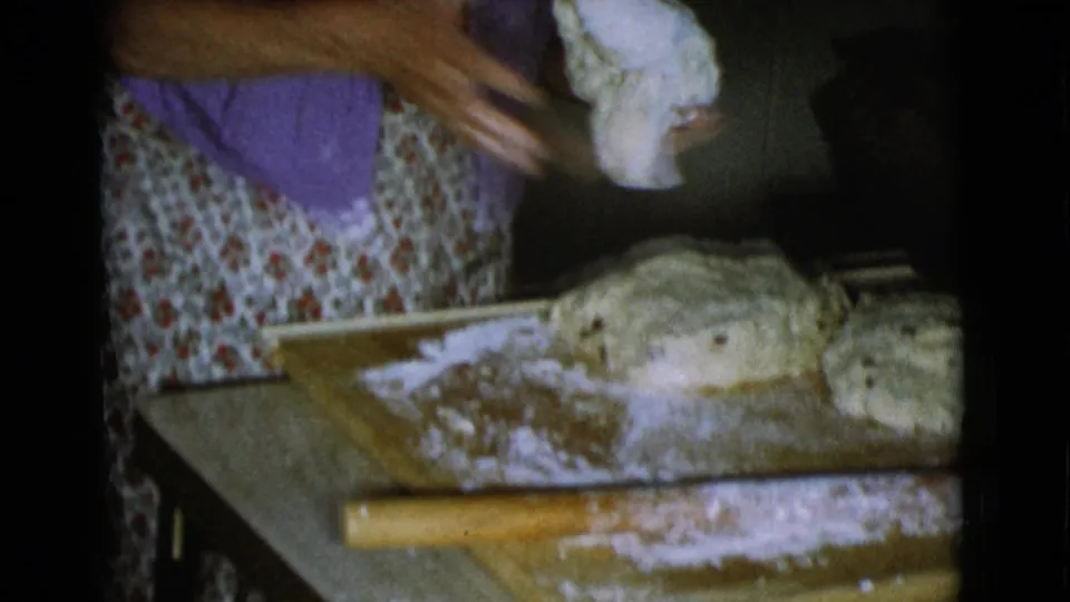 The woman holds dough above a floured board, with rounded dough portions and a rolling pin visible in the kitchen workspace.