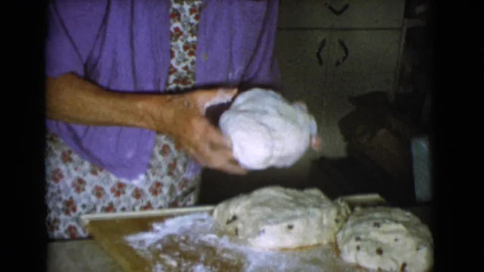 A woman in a purple shirt holds a rounded piece of dough beside other dough portions on a floured board in a private kitchen.