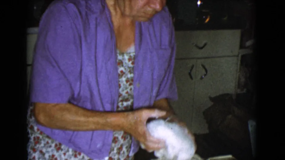 The woman continues shaping dough by hand in the 1968 Cottonwood kitchen, with cabinets and dark interior space behind her.