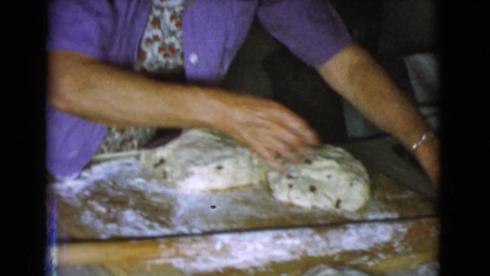 An older woman in apron and house shirt reaches across two shaped dough pieces on a floured work surface in a private kitchen.