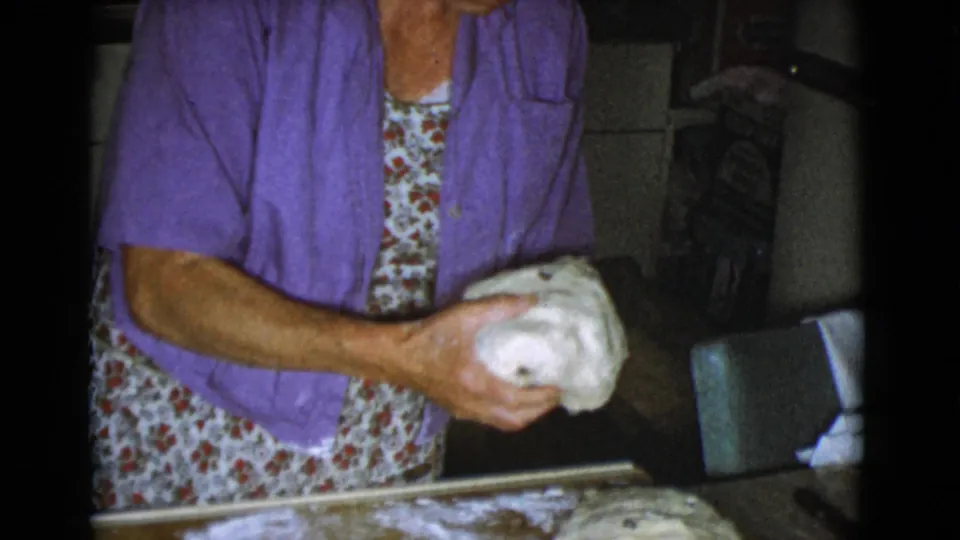 The woman continues shaping dough in a private kitchen, with flour on the board and kitchen furniture visible behind her.
