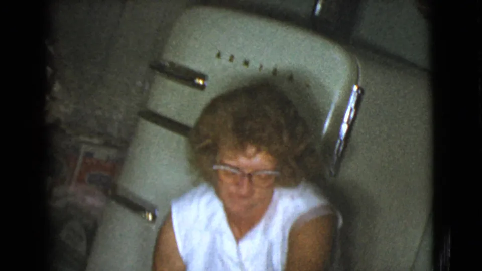An older woman wearing glasses and a white sleeveless top sits near a rounded refrigerator in the Cottonwood kitchen scene.