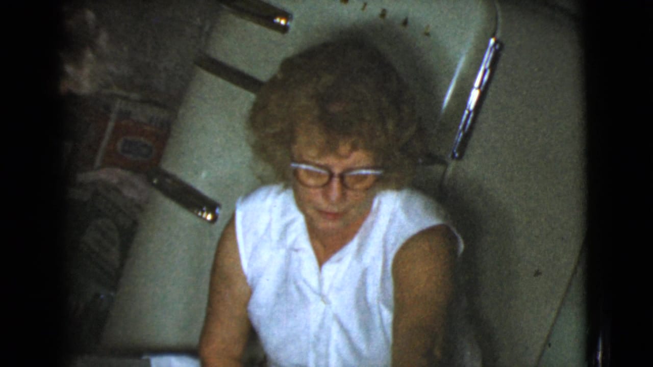 Older woman in glasses seated in a kitchen, seen from above in grainy film.