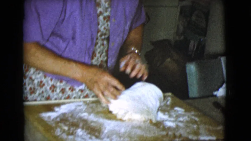 A woman in a purple shirt and floral apron kneads dough on a floured wooden board in 1968 Cottonwood home footage.