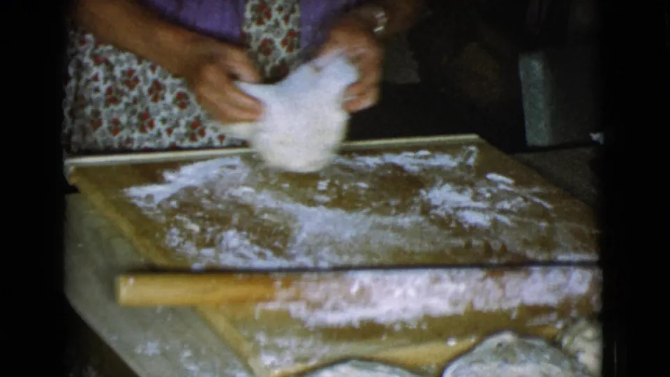 Close home-movie view of hands handling dough over a floured wooden board, with a rolling pin at the edge of the frame.
