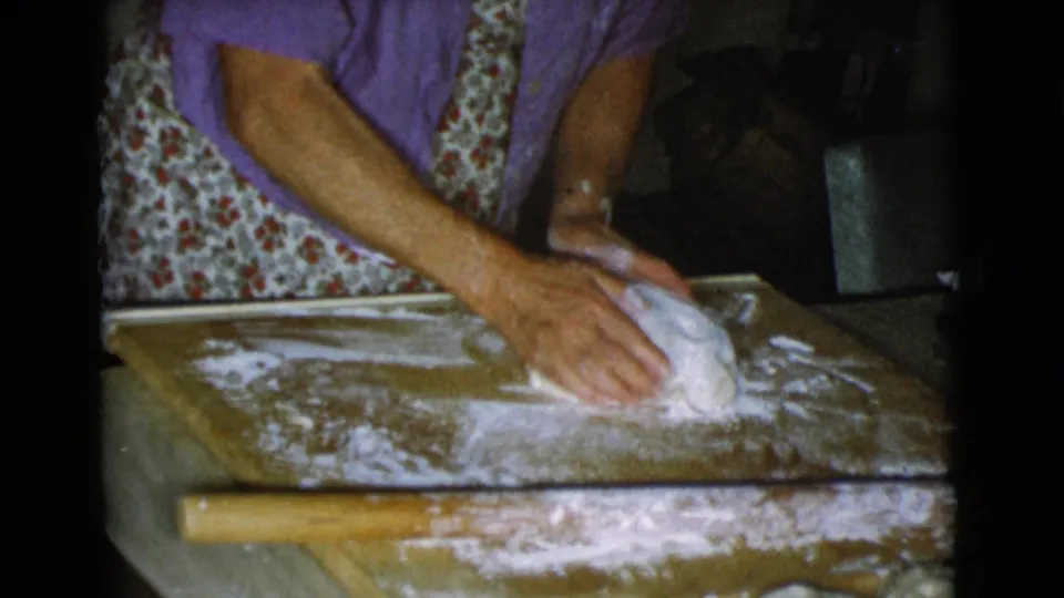 A woman presses dough across a floured wooden board, with a rolling pin visible in the foreground of the kitchen scene.