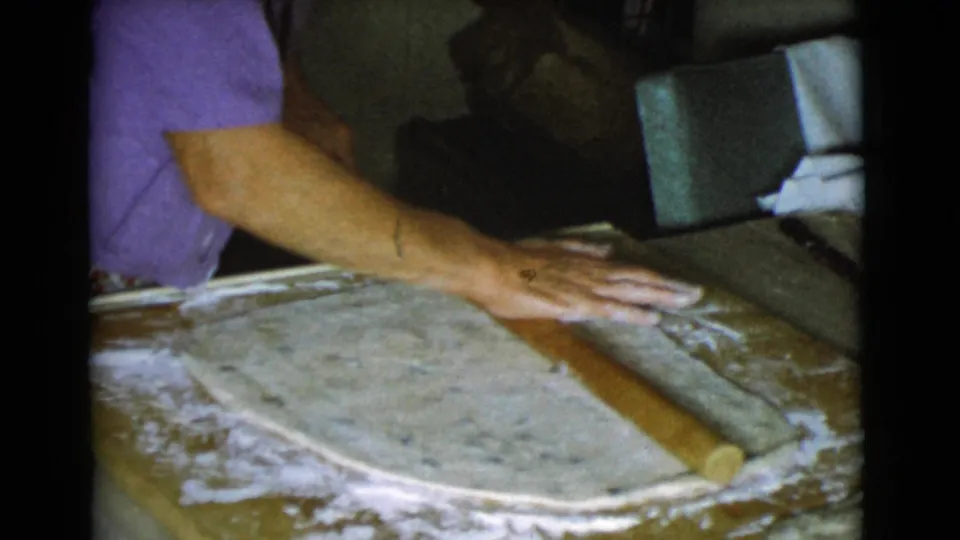 A woman continues rolling dough with a brown rolling pin in a private Cottonwood, Arizona kitchen, with chair upholstery in the background.