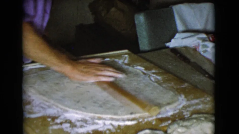 Close 8mm view of a woman's hand rolling dough on a floured surface, with a darker kitchen background visible behind the work area.