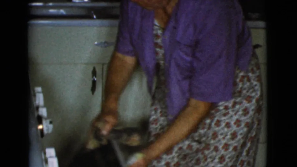 A woman in a purple shirt and floral apron bends over the kitchen work area near a pale appliance or cabinet in 1968 home-movie footage.