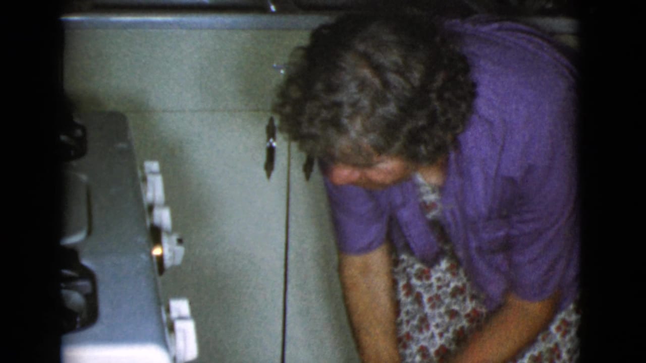 Older woman bends near a vintage oven in a small kitchen.