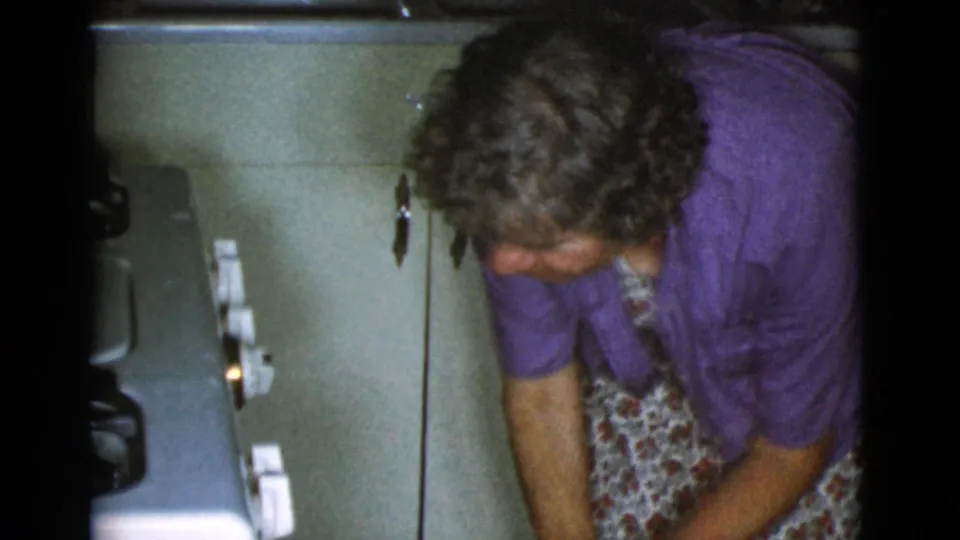 Older woman in a purple shirt and floral apron bends beside a vintage oven in the Cottonwood, Arizona 1968 home-movie scene.