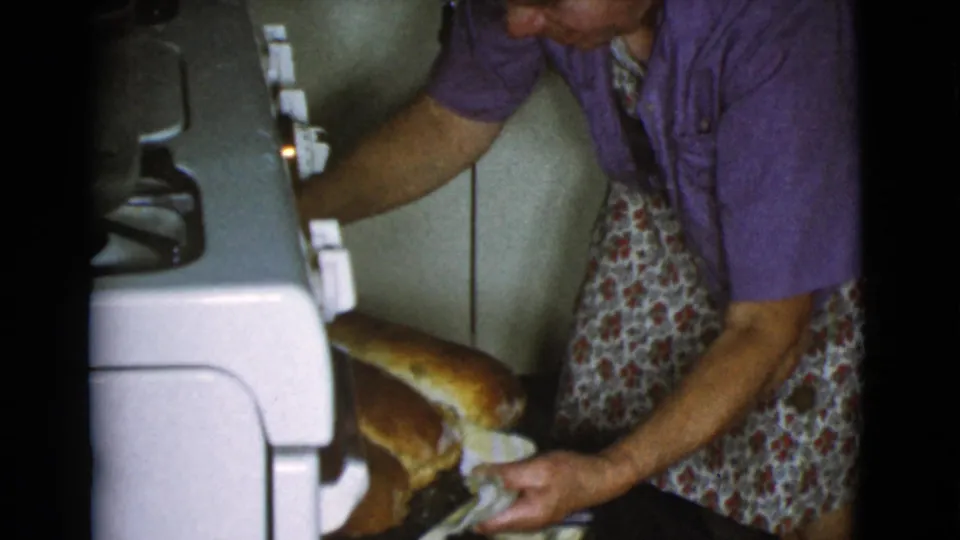 The woman in a floral apron pulls golden baked loaves from the vintage oven in the 1968 Cottonwood home kitchen footage.