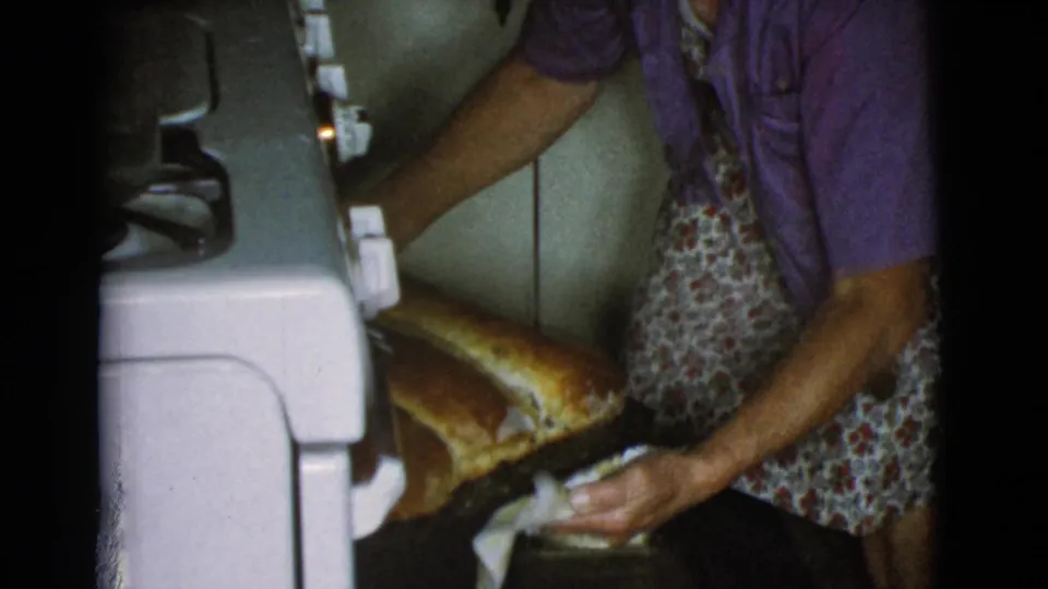 In a 1968 Cottonwood, Arizona home kitchen, a woman in a purple shirt and floral apron pulls large bread loaves from a white oven.
