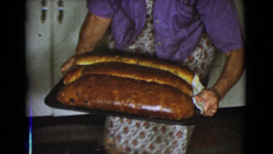 A woman in a floral apron holds a baking tray with three large golden-brown loaves inside a 1968 Cottonwood home kitchen.