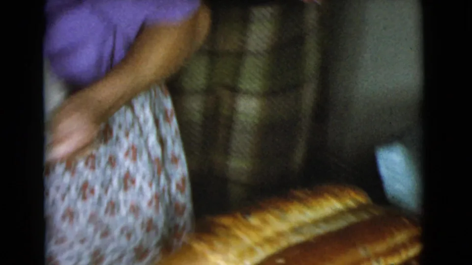 The woman moves past the tray of bread loaves in a dim 1968 home kitchen, with cabinets and household surfaces visible.
