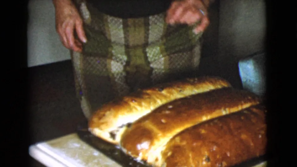 Two long homemade bread loaves rest on a counter as an adult woman in a plaid skirt stands nearby in the 1968 Cottonwood kitchen.