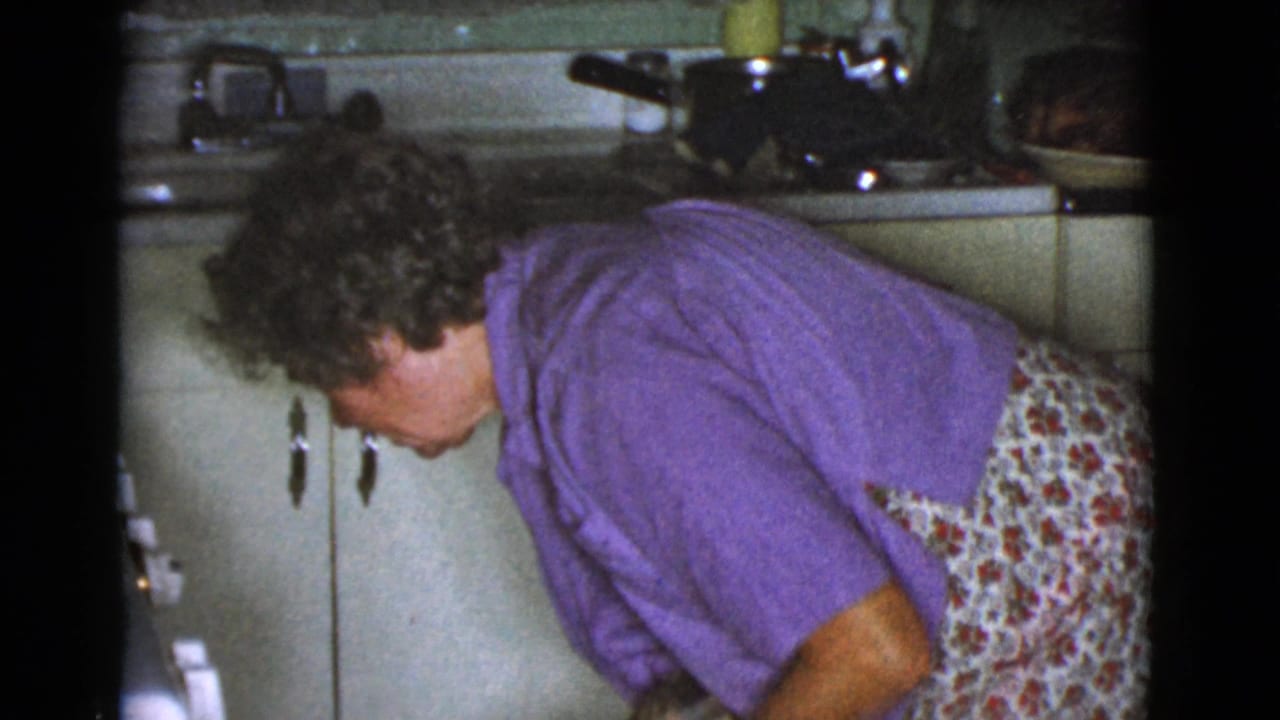Woman in a purple shirt bends over near a kitchen sink and cabinets.
