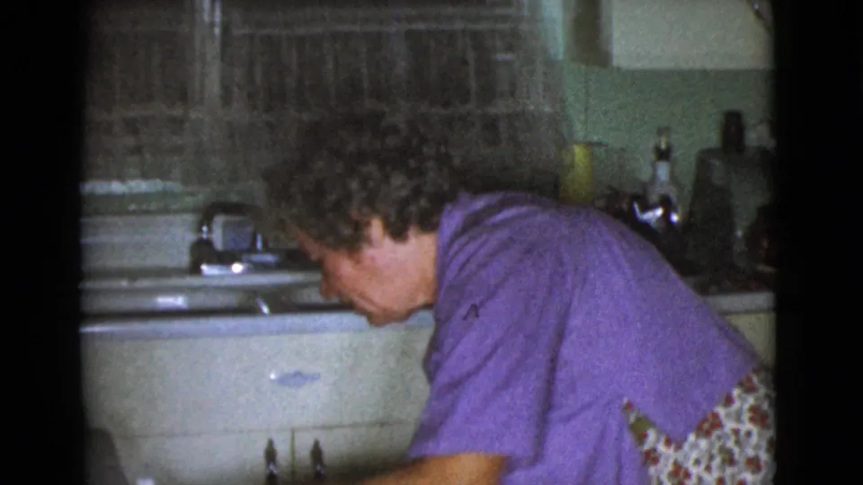 A woman in a purple shirt leans over a kitchen work area, with metal sink fixtures, pale cabinets, and patterned curtains visible.