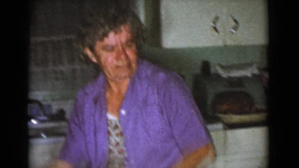The woman stands more upright in the kitchen, wearing a purple shirt over a floral garment with cabinets and counter items behind her.