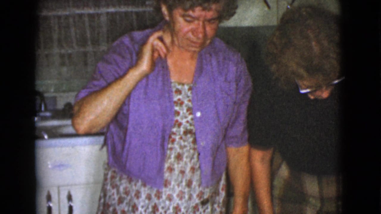 Woman in purple cardigan stands in a kitchen beside another adult bending down.
