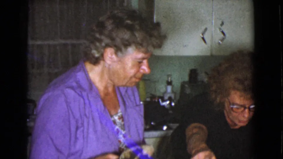 1968 home-movie frame showing two women in a modest kitchen, with cabinets and countertop items visible.
