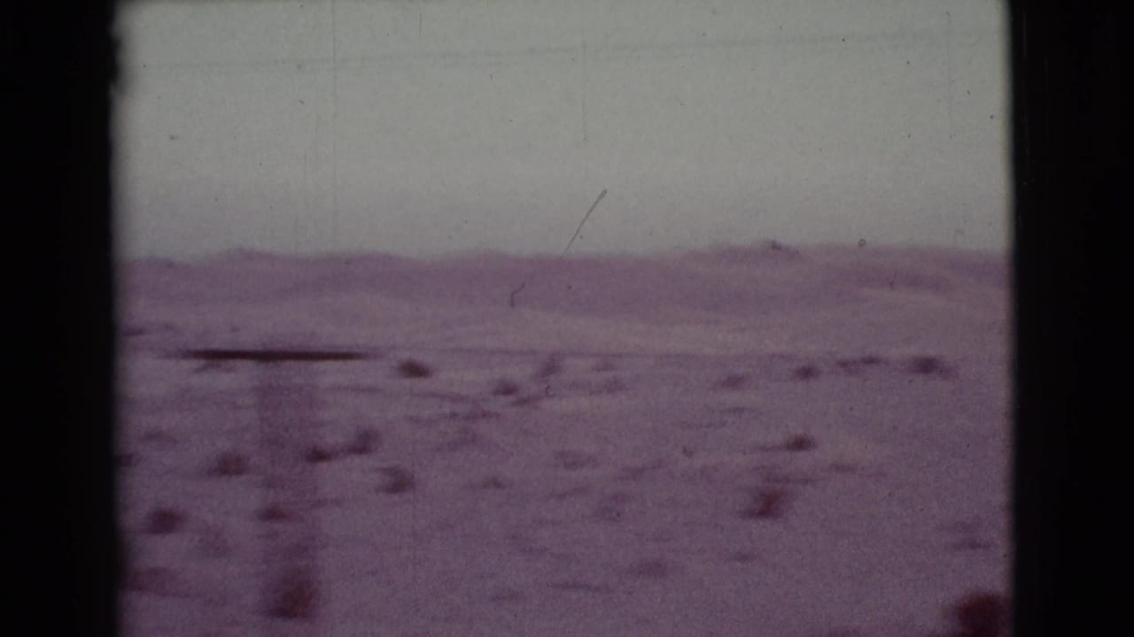 Open desert landscape with distant hills, sparse shrubs, and film-frame borders.