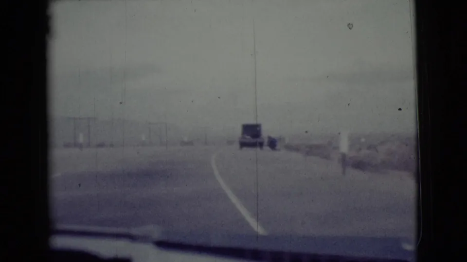 1968 Arizona 8mm windshield view of a paved highway, dashboard edge, roadside scrub, and a sedan ahead in the distance.