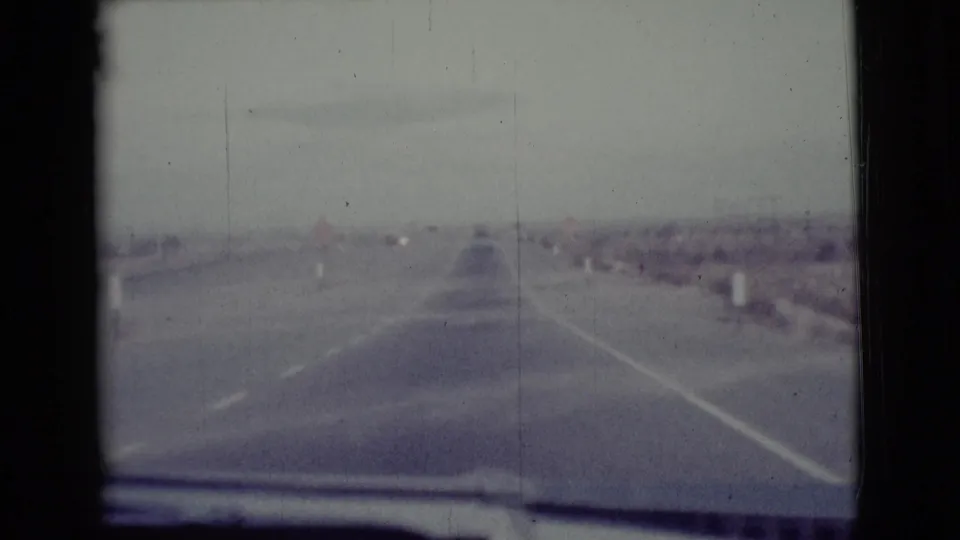 A 1968 Cottonwood-area road view from inside a moving car, with lane markings, roadside posts, and open arid ground ahead.