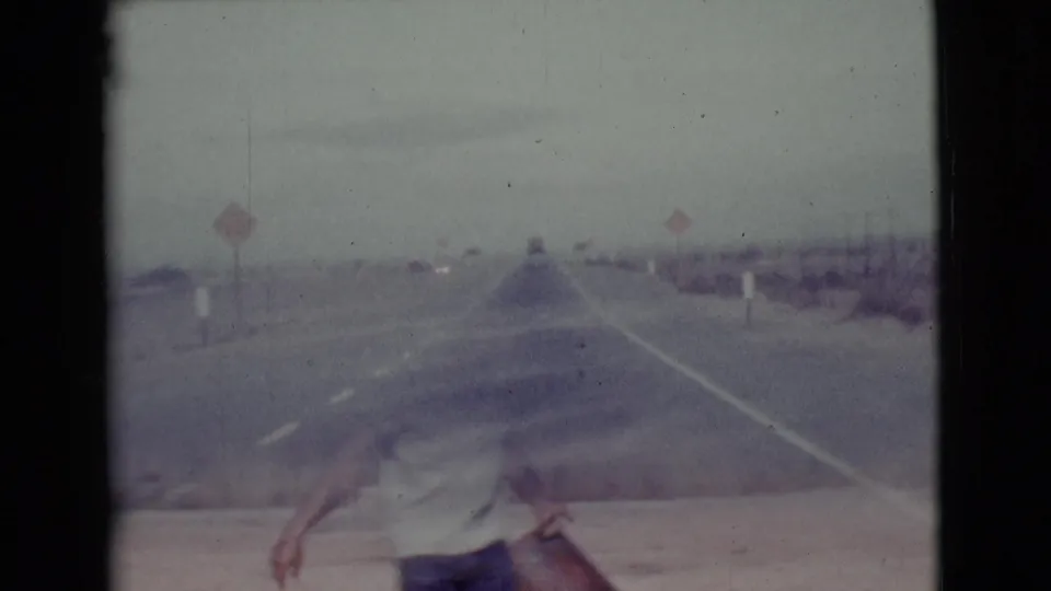 Faded 8mm footage shows a hazy Arizona road scene with roadside signs and scrubland as seen from a moving vehicle.