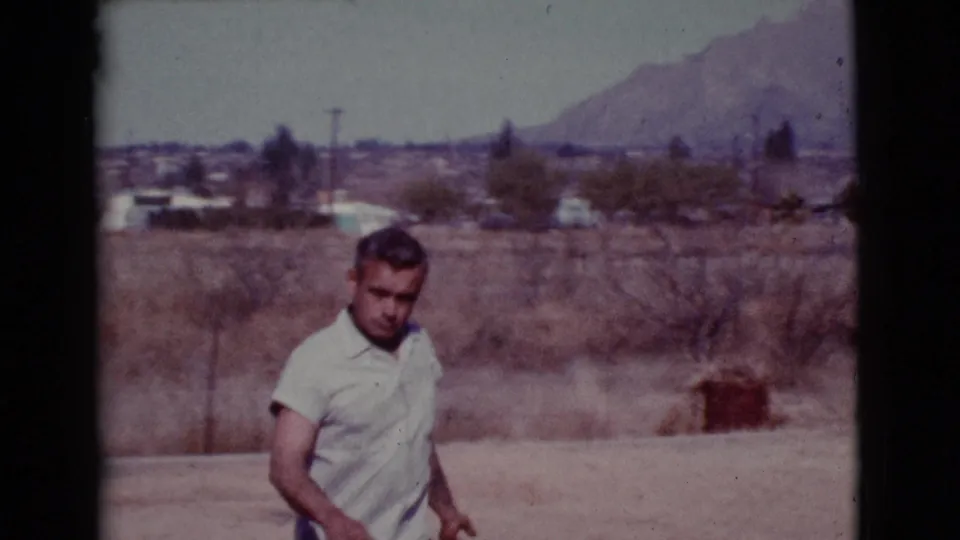 In Cottonwood, Arizona, an adult man in a light short-sleeve shirt walks across a dusty field with low mountains and scattered buildings behind him.