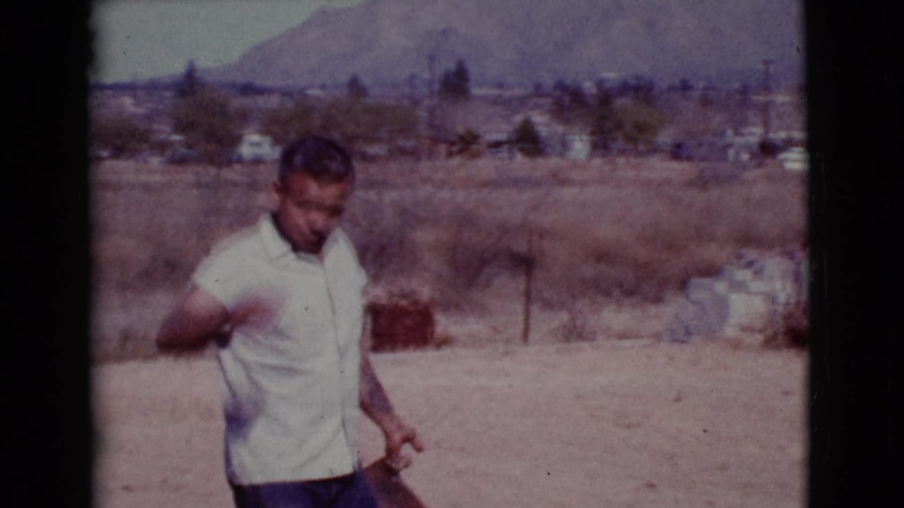 Man in a light shirt holding a shovel in a dry field with hills behind him.