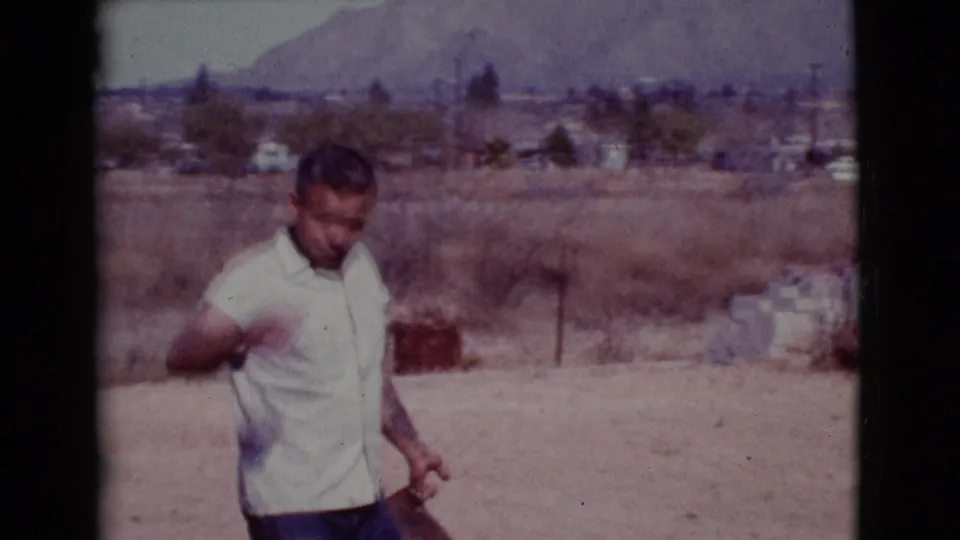 In Cottonwood, Arizona, 1968, a man stands in a dry field holding a shovel, with low buildings and distant hills in the background.
