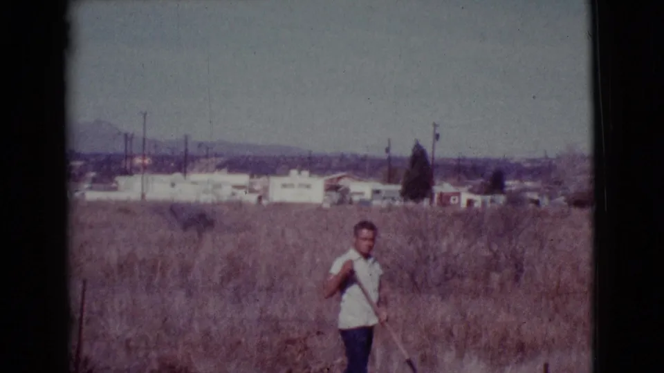 A man walks through tall dry grass in Cottonwood, Arizona, with a shovel and low suburban buildings visible beyond the field.