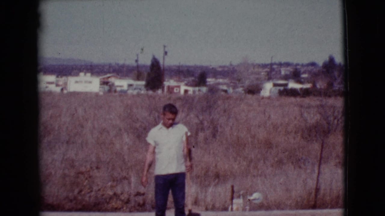 Man in a white shirt stands in a dry field with buildings and hills behind him.