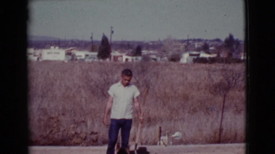 A man in a white shirt looks toward the ground in a dry Cottonwood, Arizona field, with homes, poles, and hills in the background.