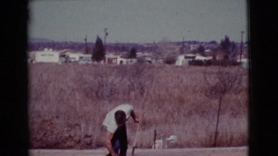 The man bends toward something near the ground in the dry Arizona field, with brush, buildings, and utility poles visible behind him.