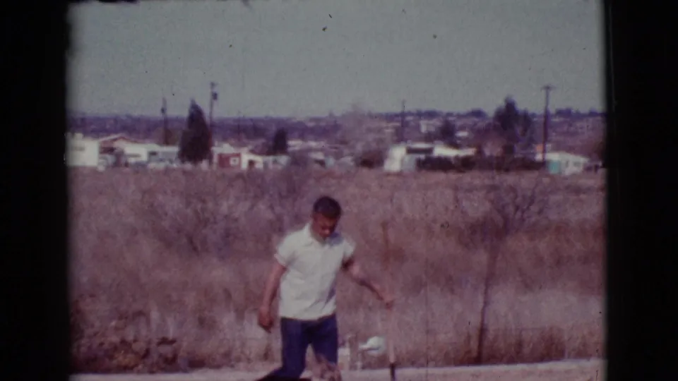 A man steps along the edge of a dry field in Cottonwood, Arizona, with a long-handled tool, pale buildings, utility poles, and hills behind him.