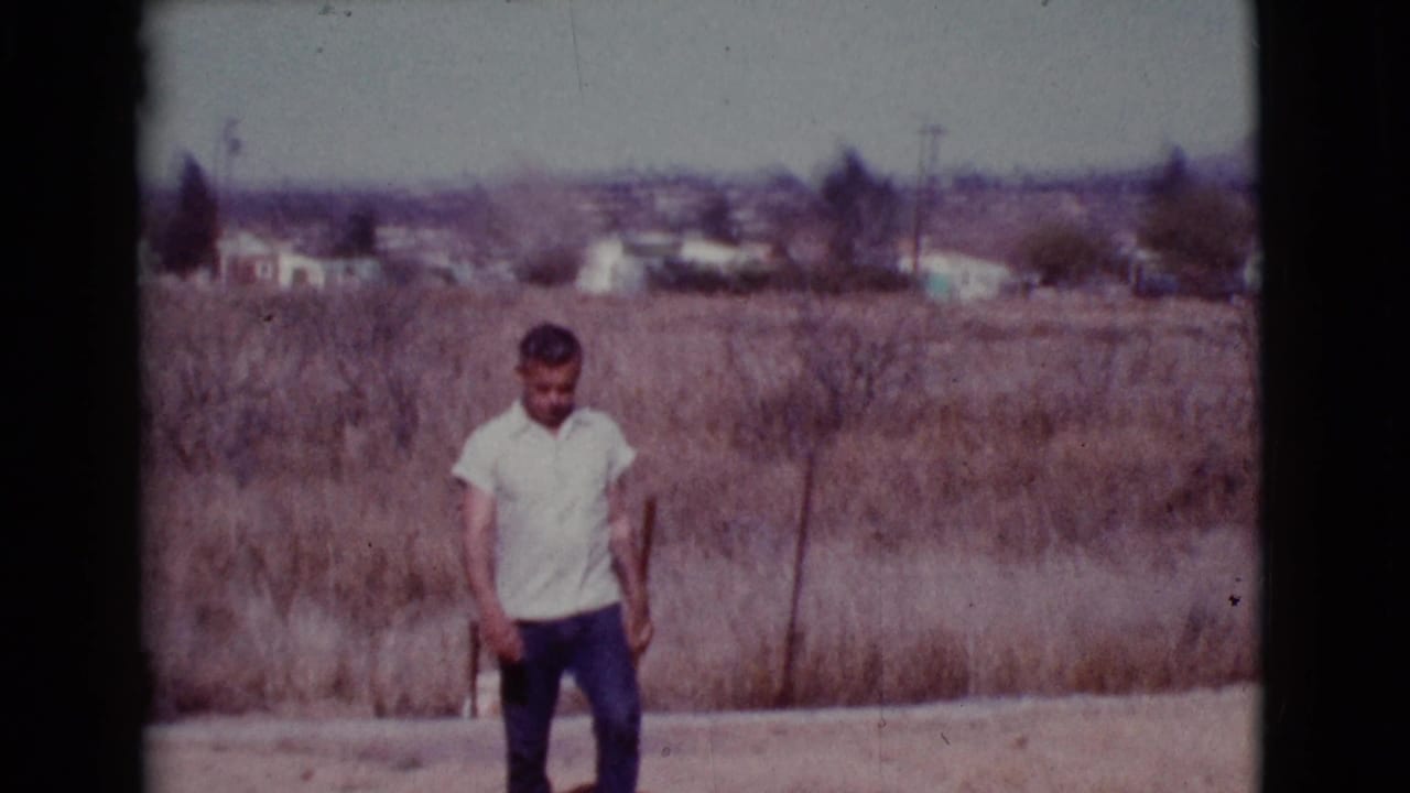 Man in a light shirt stands in a dry field with distant houses behind him.