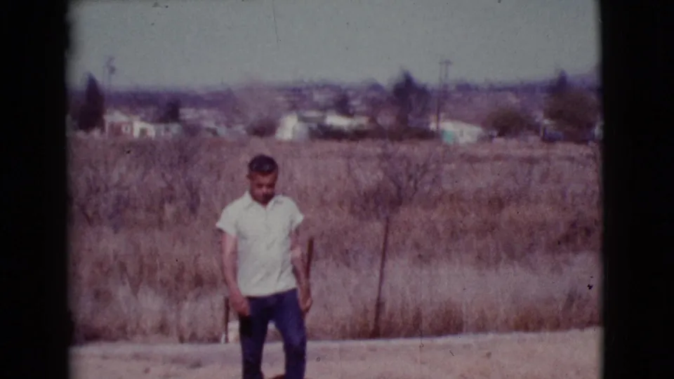In Cottonwood, Arizona, a man in a light shirt stands on dry open ground with brush and distant houses visible in this 1968 8mm frame.