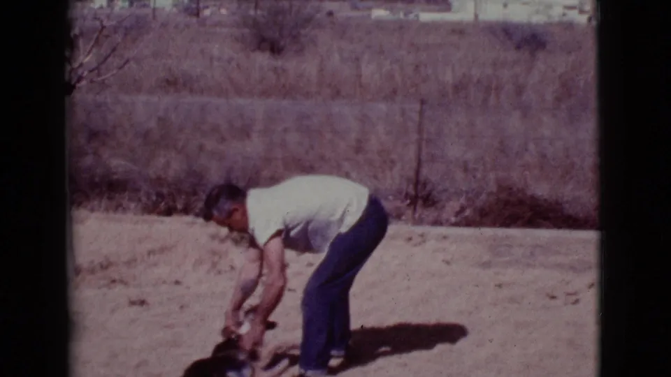 A man in casual clothes crouches toward the small dog on sunlit dirt in the 1968 home-movie scene.