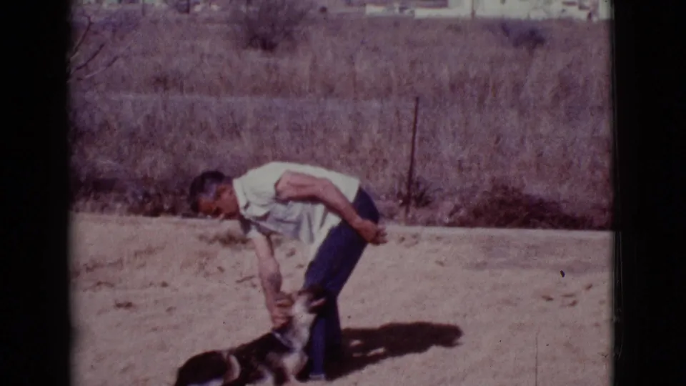The man bends over the small dog near his feet, with sparse brush and strong sunlight behind them.