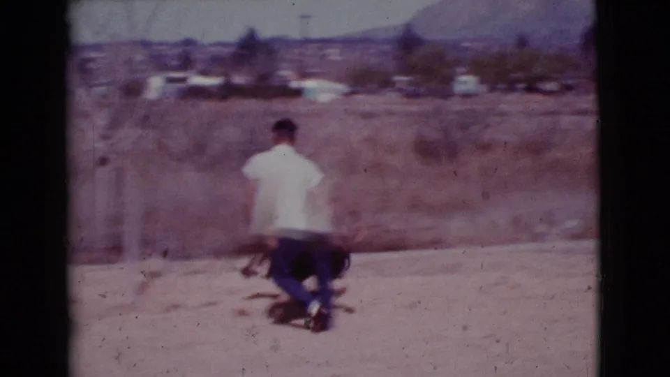 The man, seen from behind in a dry Cottonwood field, moves with a dark dog near sparse trees and distant houses.
