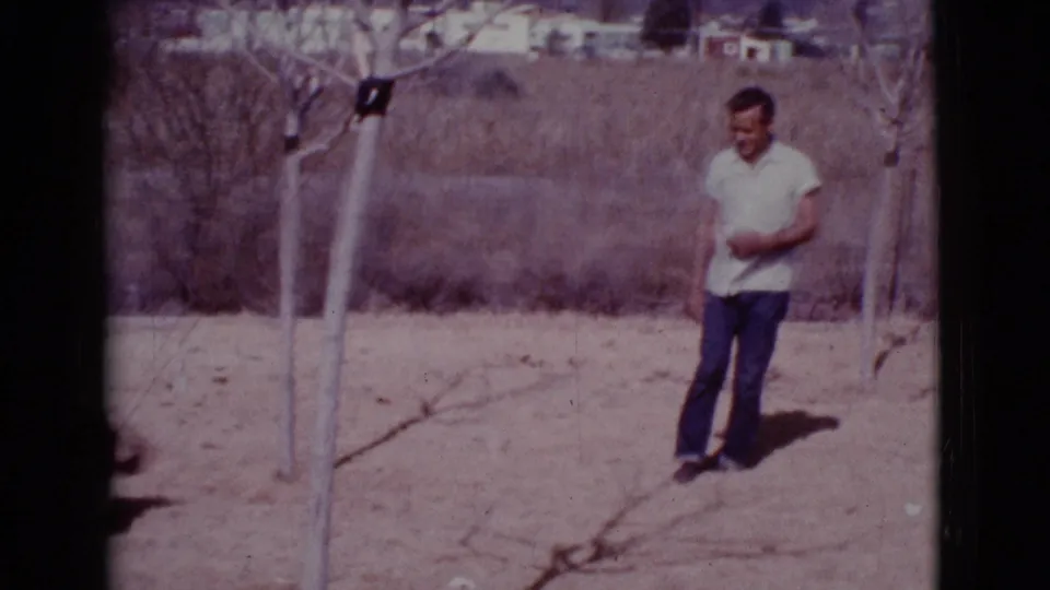 A man in a light shirt stands near slender trees in the dry Cottonwood, Arizona field during the 1968 home movie.