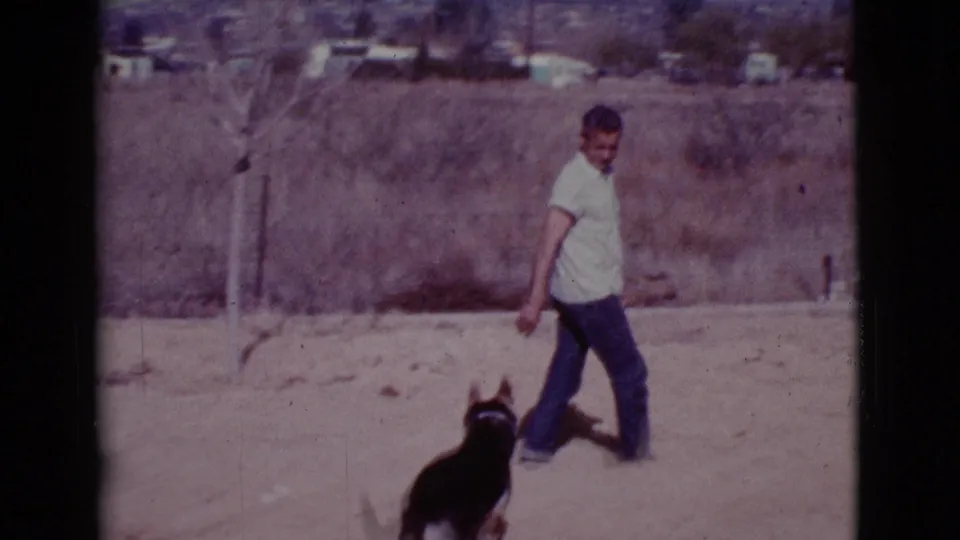 In Cottonwood, Arizona, 1968, a man in a white short-sleeve shirt walks beside a black-and-white dog on dusty ground.