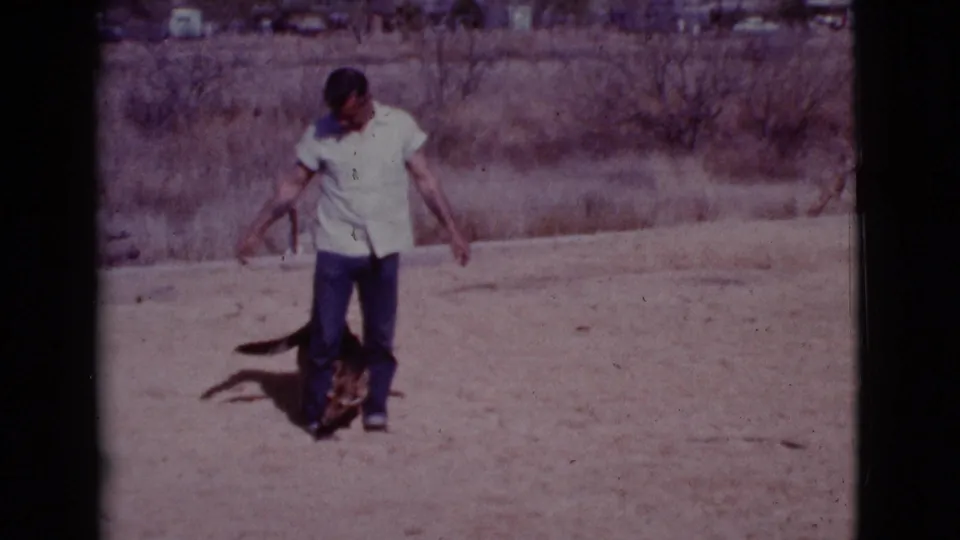 In the Cottonwood, Arizona scene, the man looks down while the dog moves close around his legs on pale dirt.