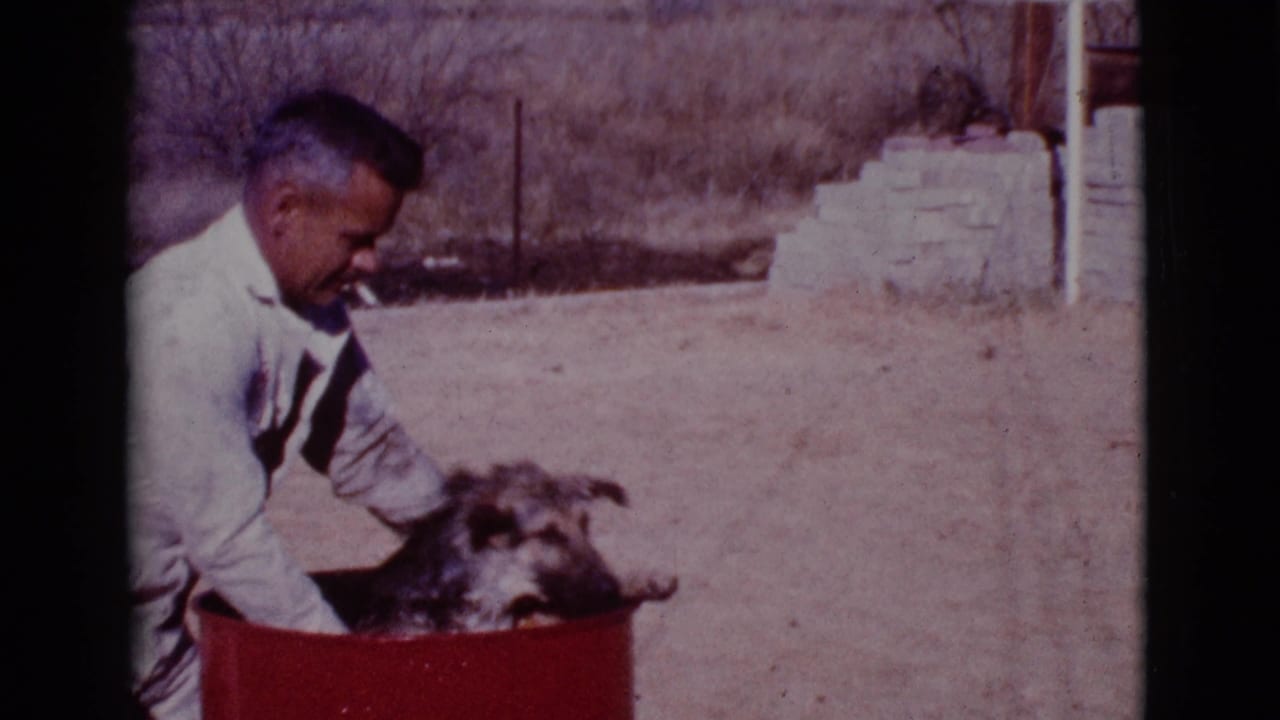 Man crouching beside a dog in a red barrel in a dry outdoor yard.